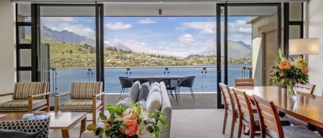 A modern room with a large sliding glass door leading to a balcony overlooking a lake and mountains. Inside, there is a dining table with chairs, a sofa, and a coffee table with a vase of flowers.