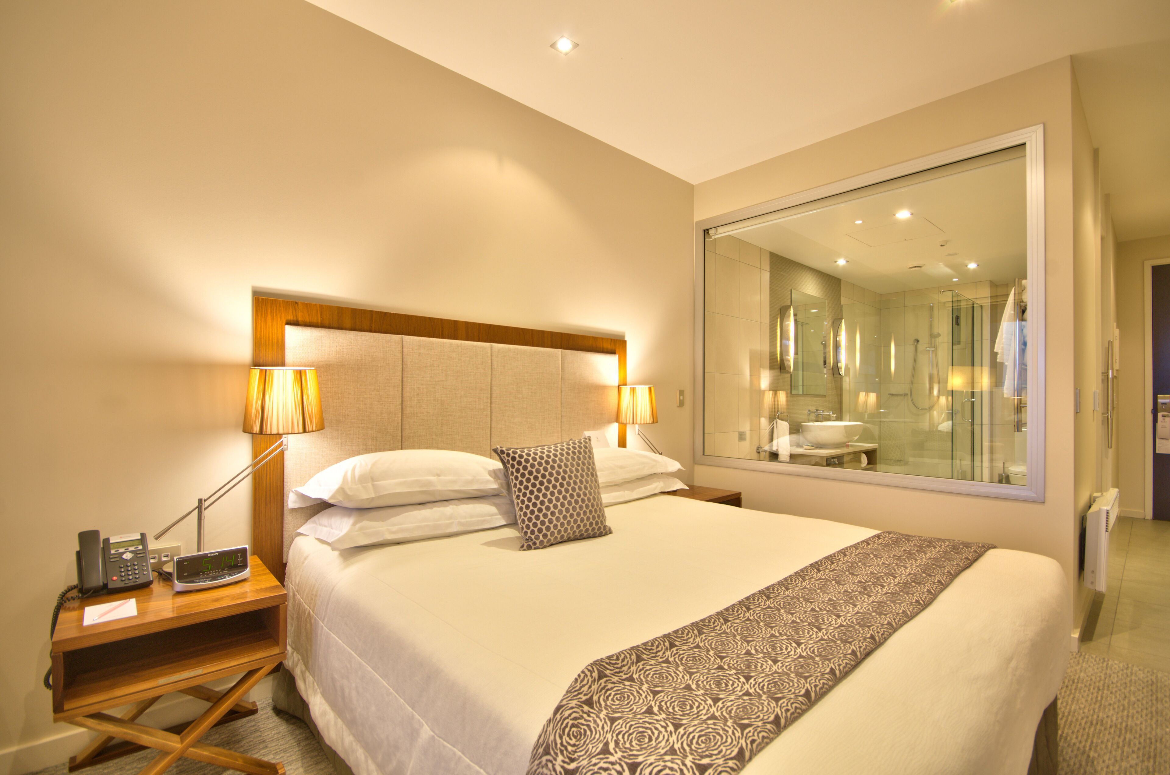 Habitación de hotel con una cama grande, lámparas de noche, un teléfono y un espejo que refleja el baño.