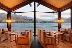 A room with a large window offering a view of a lake and mountains. There are wooden chairs and tables arranged for seating.