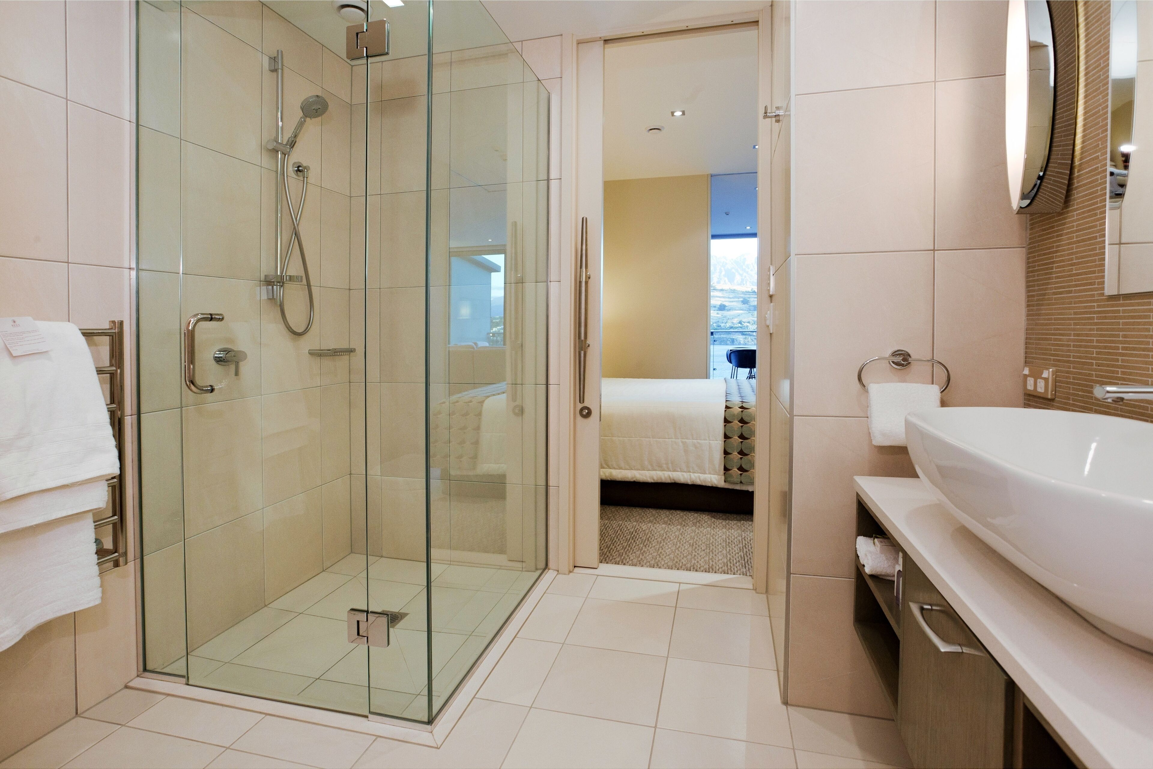 A modern bathroom with a glass-enclosed shower, a large sink, and a visible bedroom in the background.