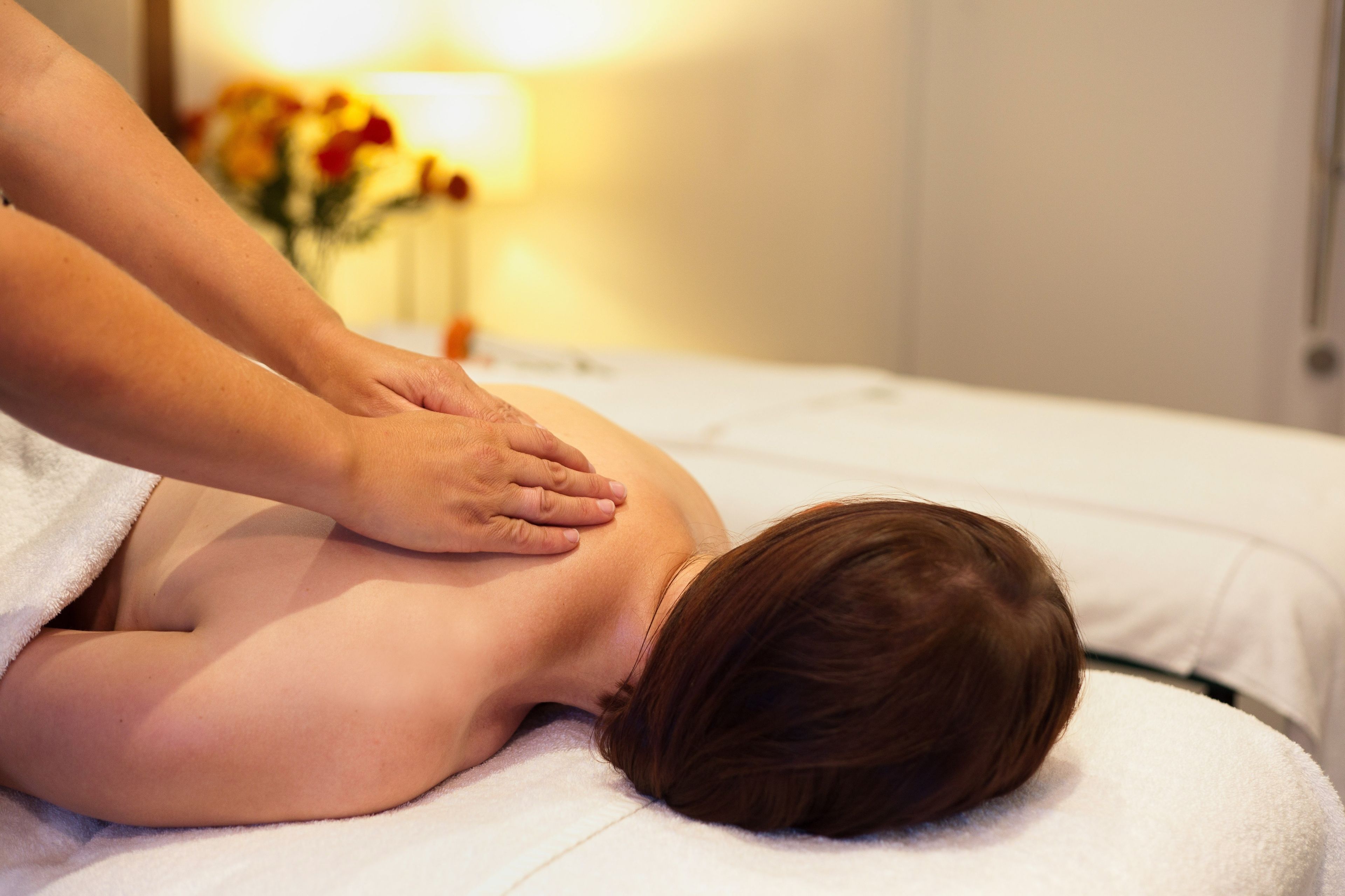 A person receiving a back massage on a bed in a room with a lamp and flowers in the background.