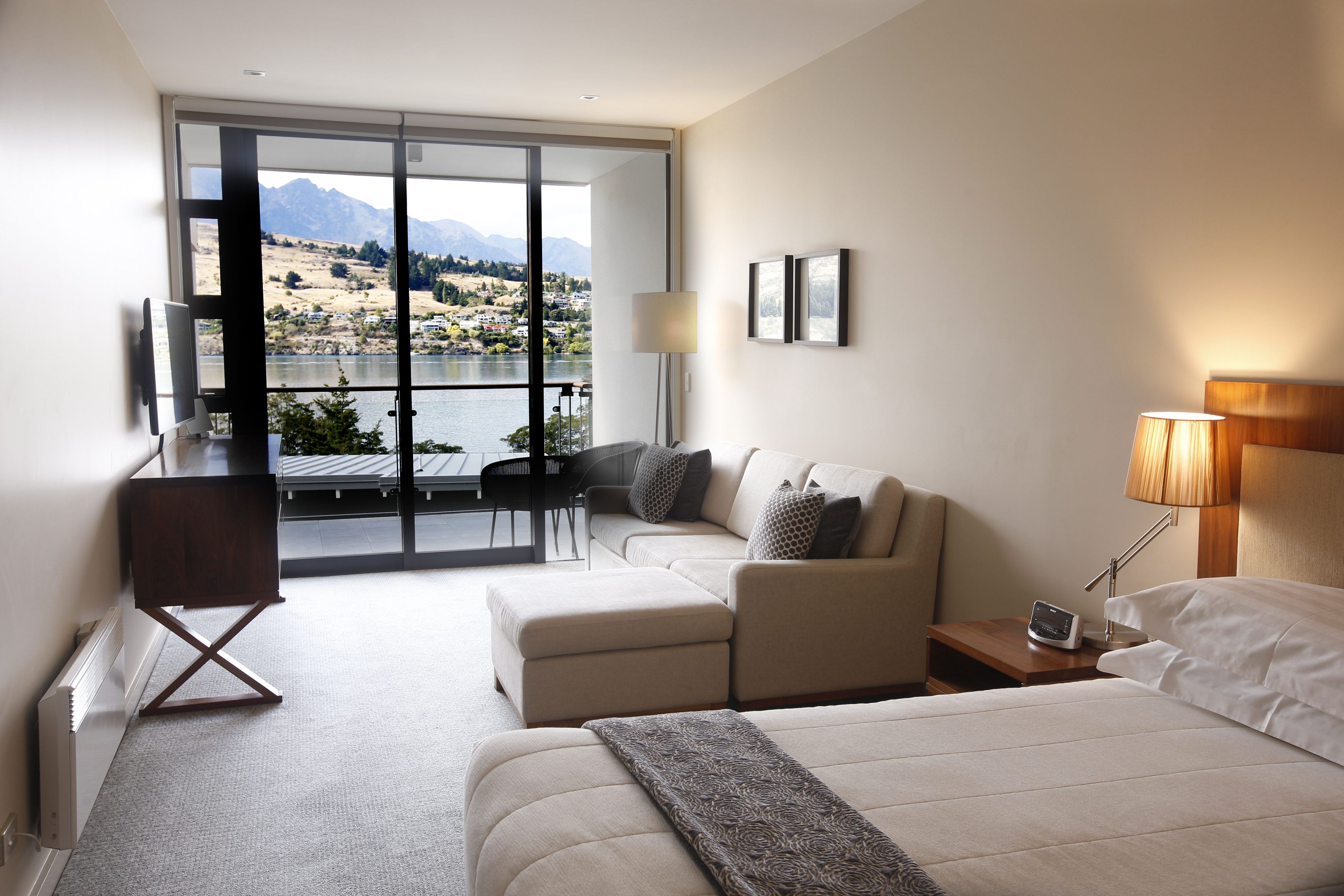 A modern hotel room with a large bed, a sofa, a desk, and a view of a lake and mountains.