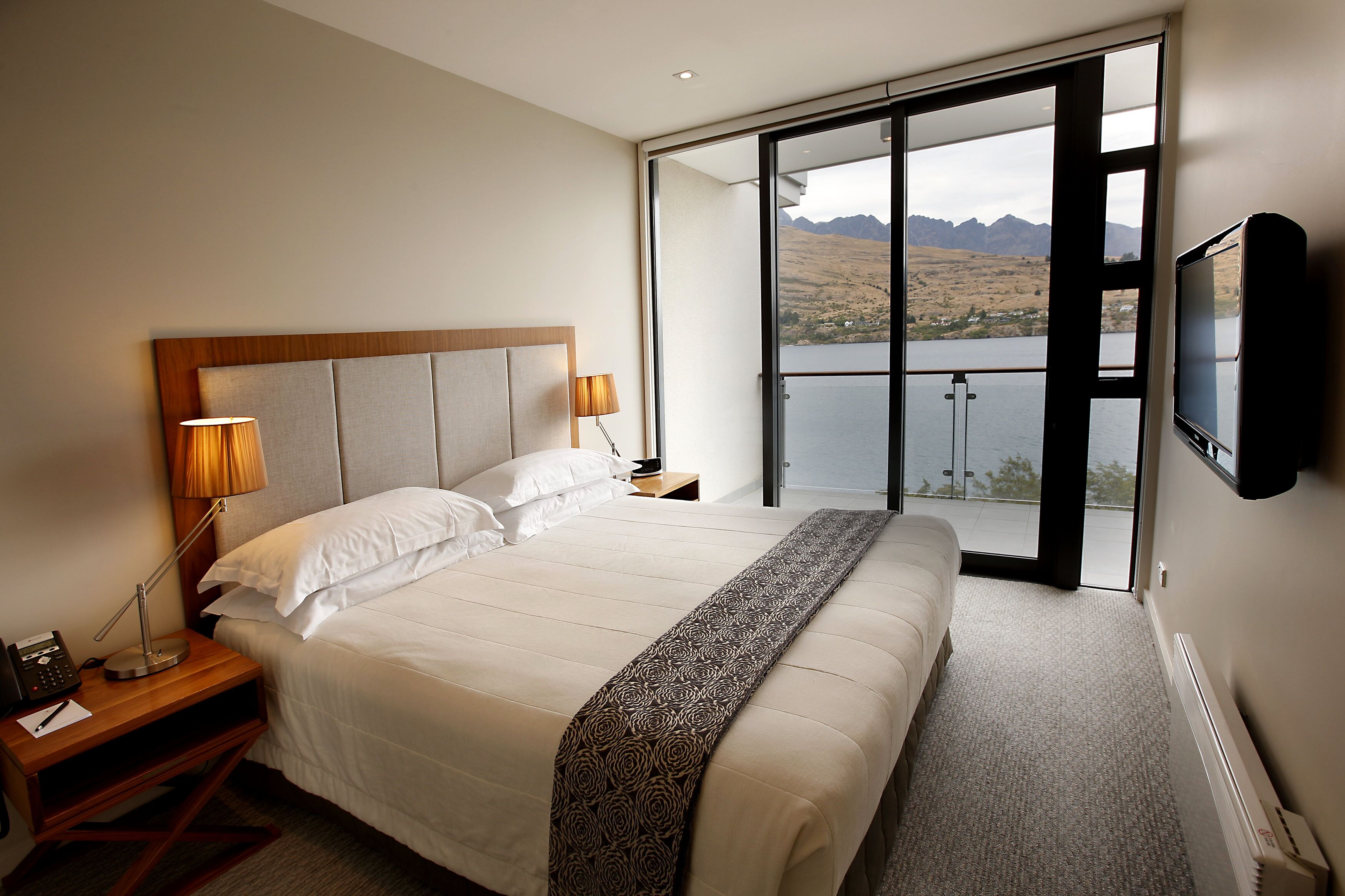 Ein Hotelzimmer mit einem großen Bett, einem an der Wand montierten Fernseher und einem Balkon mit Blick auf Berge und einen See.