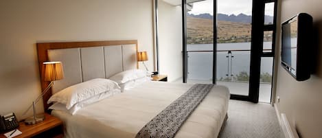 A hotel room with a large bed, a TV mounted on the wall, and a balcony with a view of mountains and a lake.