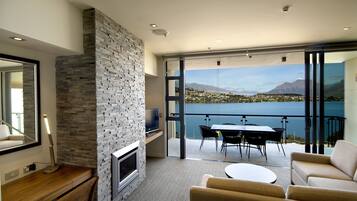 A modern living room with a stone fireplace, a flat-screen TV, and a sofa. It features a large sliding glass door leading to a balcony with a view of a lake and mountains.