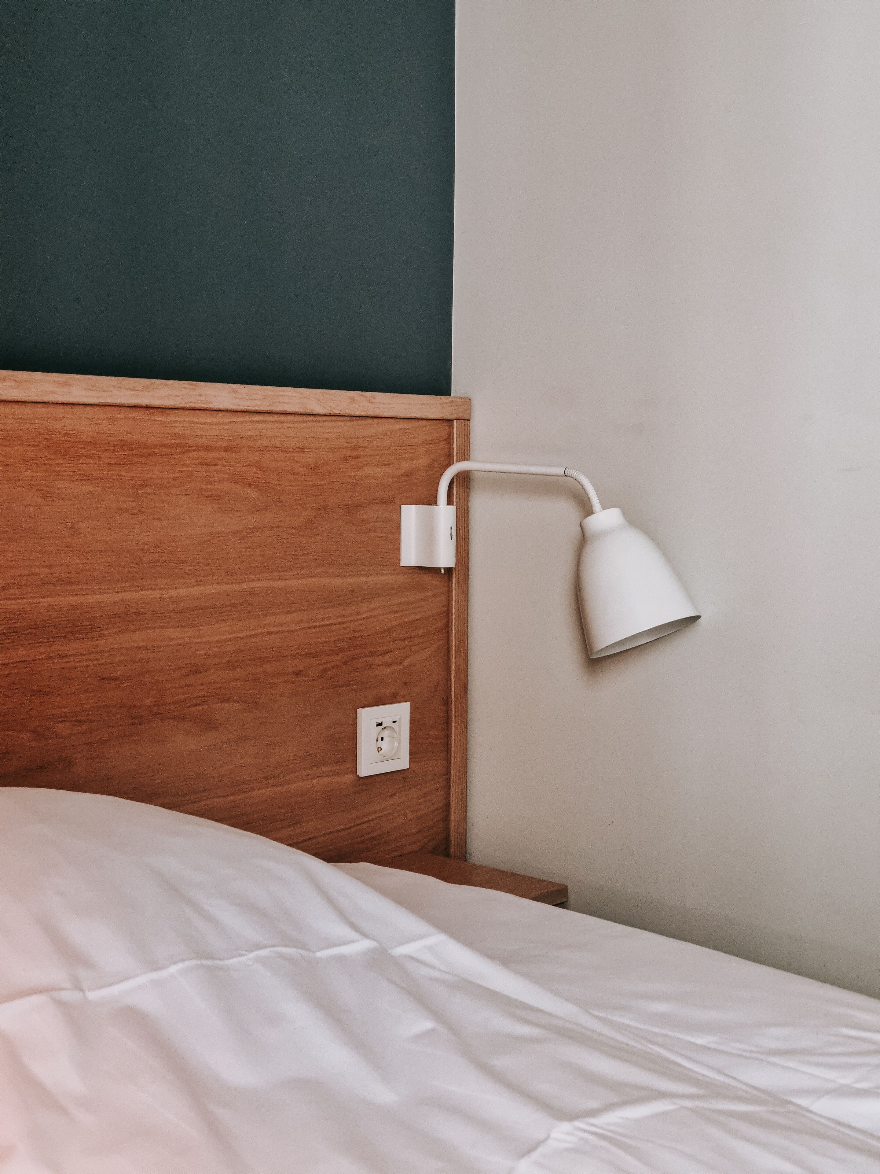 A bed with a wooden headboard, a white bedside lamp, and a power outlet on the wall.