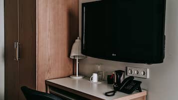 A hotel room with a wooden wardrobe, a desk with a lamp, a flat-screen TV, a coffee maker, and a telephone.