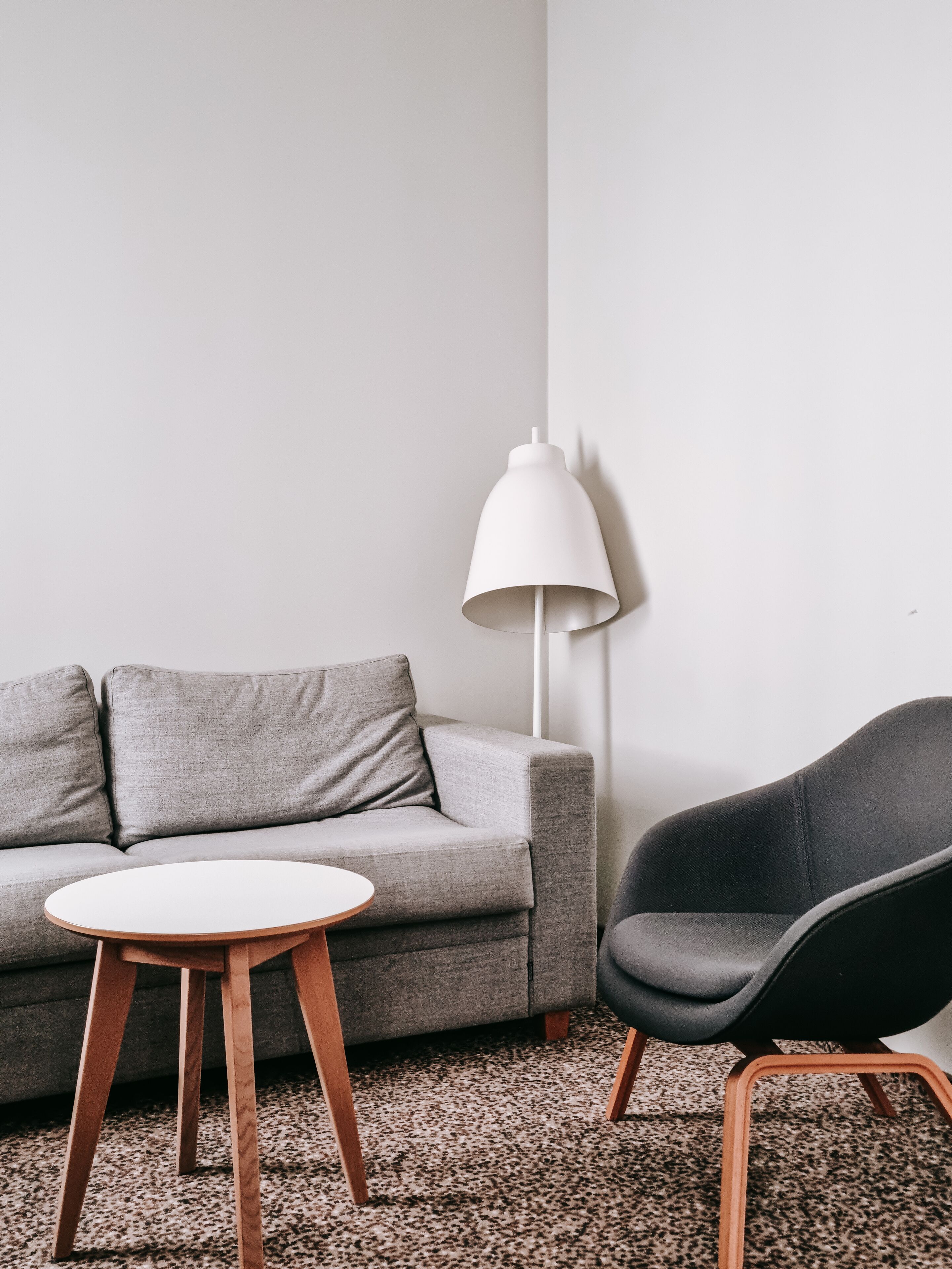 A living room with a grey sofa, a round wooden table, a black chair, and a white floor lamp.