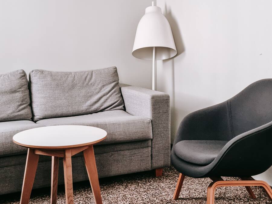 A living room with a grey sofa, a round wooden table, a black chair, and a white floor lamp.