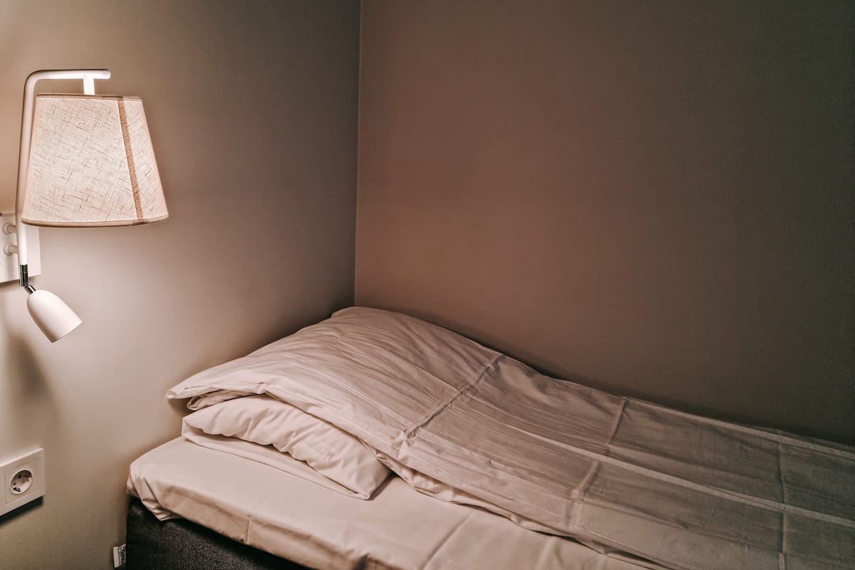 A small, neatly made bed with a plain wall and a single bedside lamp.