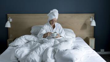 Una persona con una bata blanca sentada en una cama con ropa de cama blanca, sosteniendo una taza. Hay dos mesitas de noche con lámparas, un cabecero de madera y una pared oscura al fondo.
