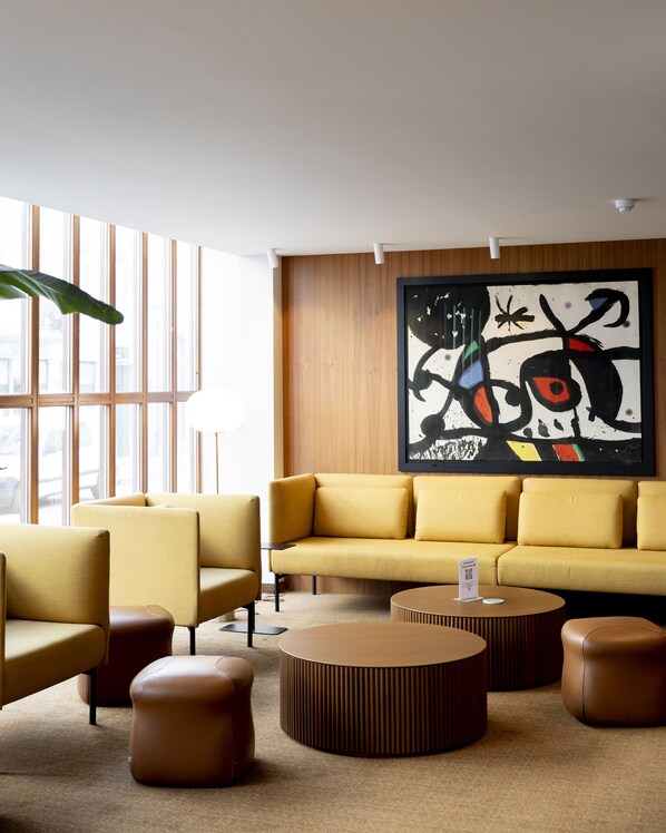A modern living room with a yellow sofa, two armchairs, and a coffee table. A large abstract painting is on the wall, and there is a window with vertical blinds. - Hotel Noreg (Ålesund)