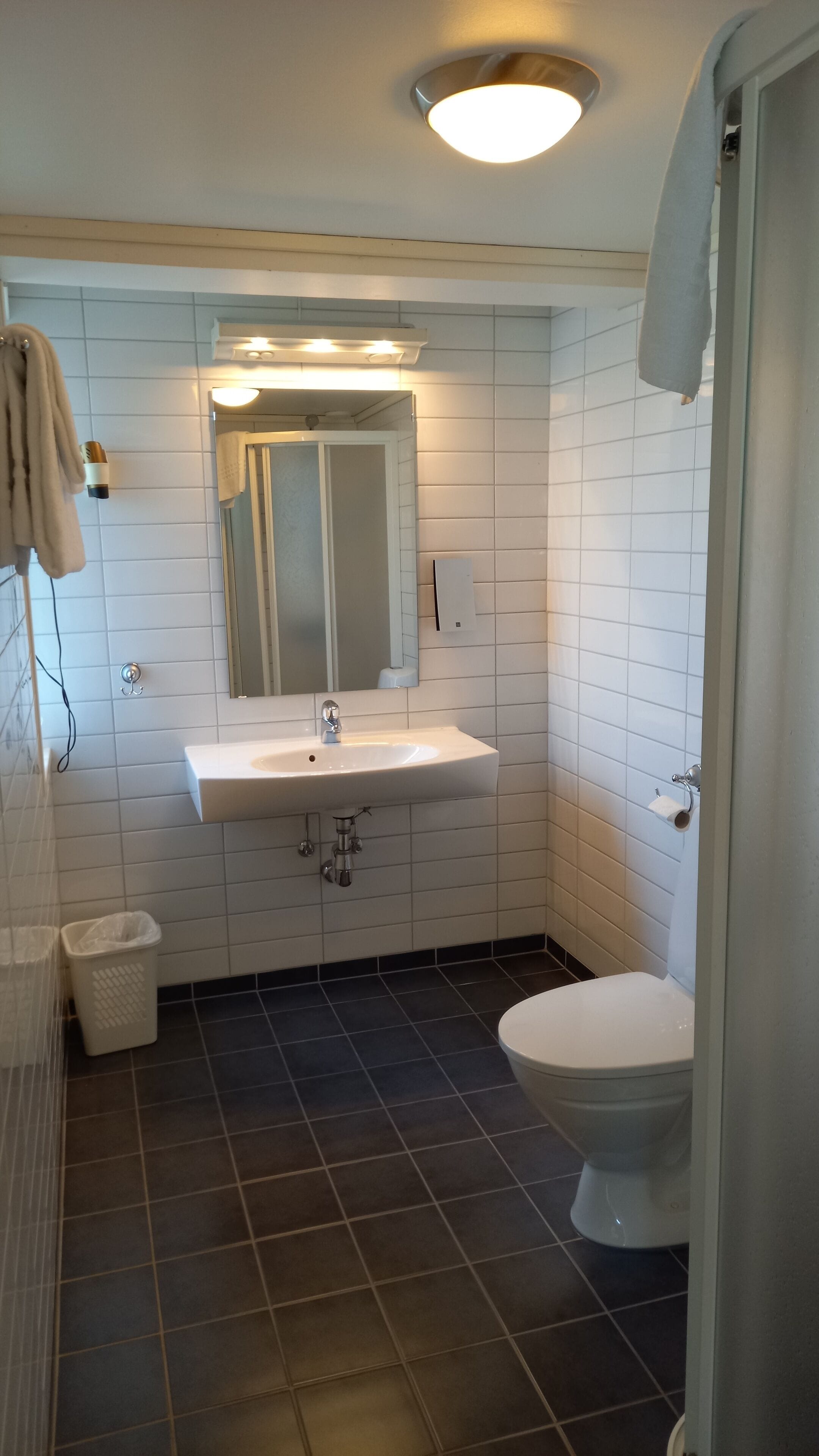 A bathroom with a white sink, mirror, and toilet. There is a towel hanging on the wall and a trash bin.