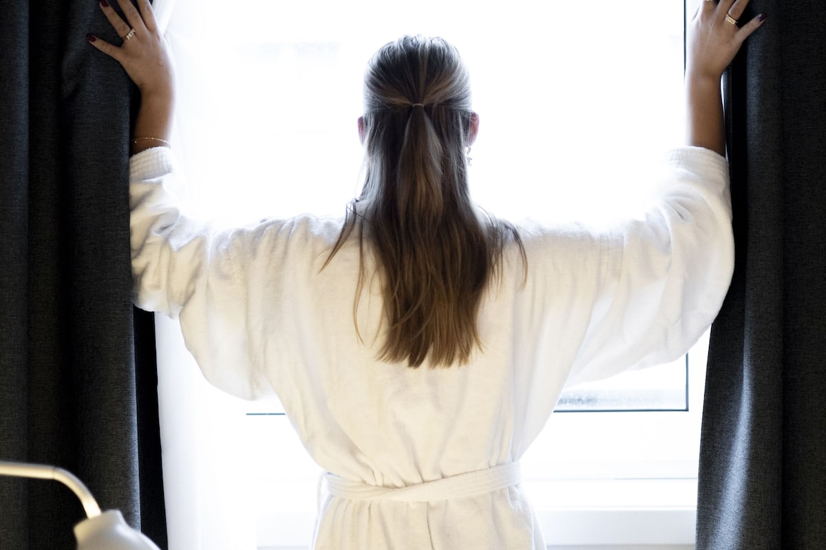A person in a white robe standing by a window with curtains, facing away from the camera.