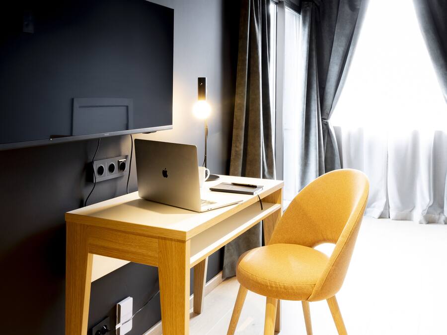 Habitació moderna amb escriptori de fusta, un ordinador portàtil, una cadira groga, televisió de pantalla plana penjada a la paret i una finestra amb cortines.