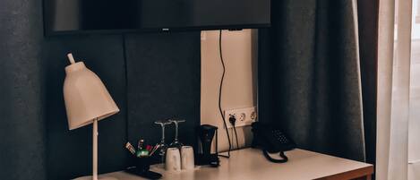 A hotel room with a desk, a chair, a TV mounted on the wall, a lamp, and a window with curtains.