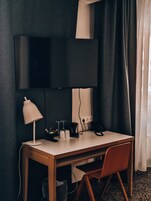Habitación de hotel con escritorio, silla, televisión en la pared, lámpara y ventana con cortinas.