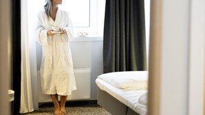 A room with a bed, a window with curtains, and a person in a robe. - Hotel Noreg (Ålesund)