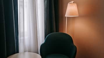 A room with a dark green armchair, a small round table, a floor lamp, and sheer curtains.