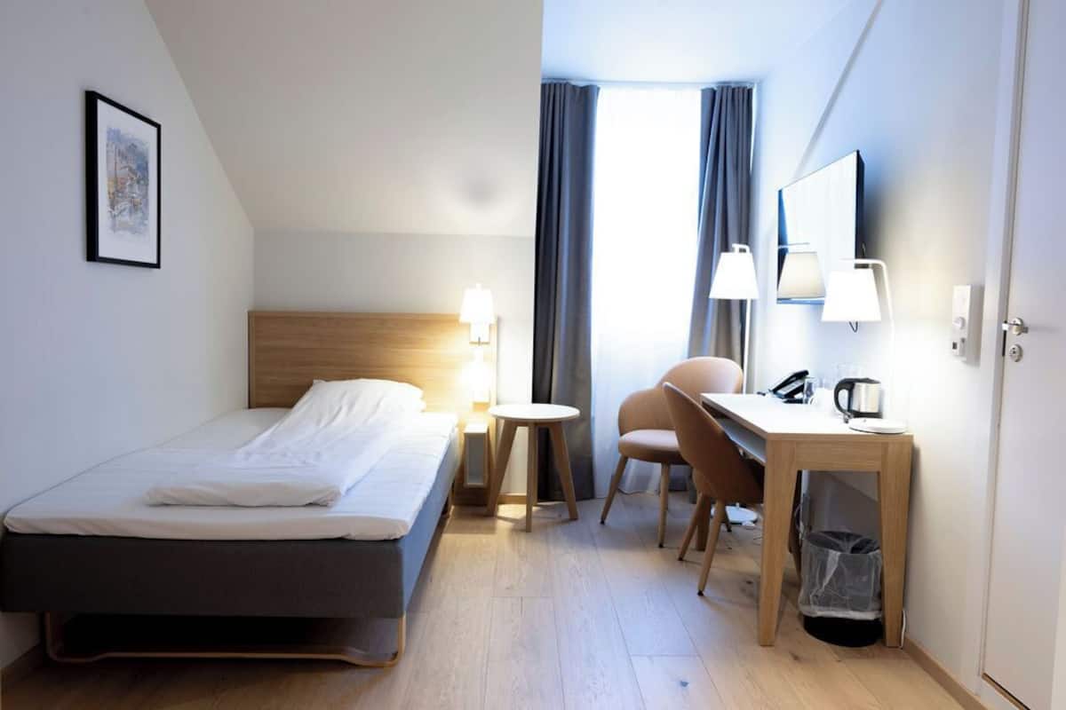 Habitación de hotel con cama, escritorio, silla, mesita, lámpara, un cuadro en la pared y ventana con cortinas.