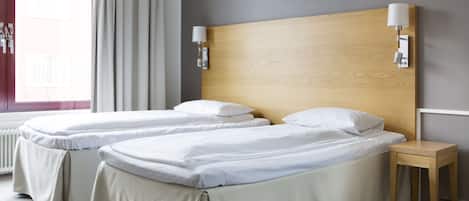 A hotel room with two single beds, a wooden headboard, bedside lamps, a small wooden nightstand, and a window with curtains.