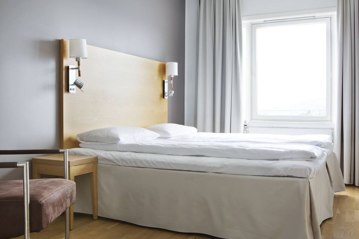 A hotel room with a bed, bedside tables, a lamp, a chair, and a window with curtains.