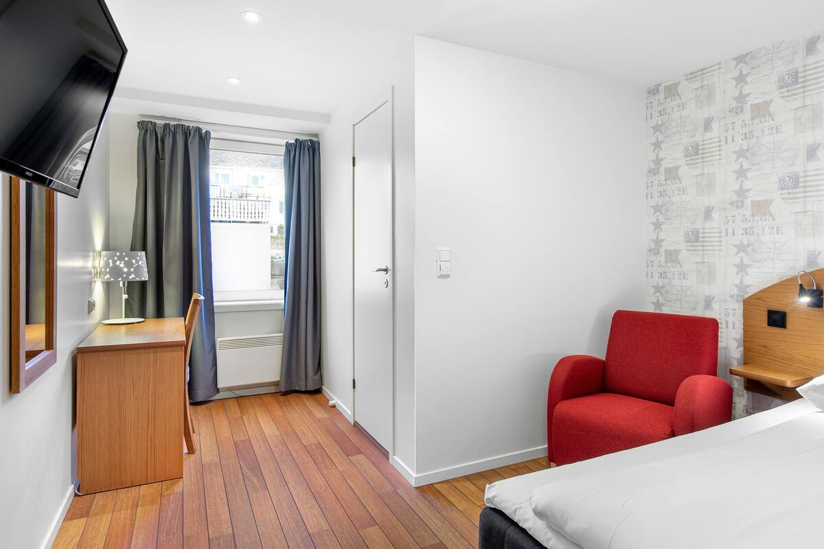 A hotel room with a wooden desk, a red chair, a bed, and a TV mounted on the wall.