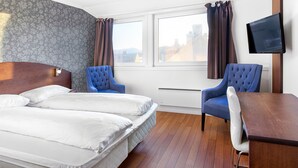 Hotellihuone, jossa on parivuode, kaksi sinistä nojatuolia, puinen työpöytä ja seinälle asennettu litteänäyttöinen televisio.