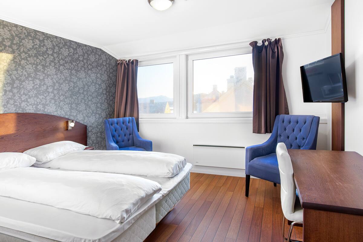 A hotel room with a double bed, two blue armchairs, a wooden desk, and a flat-screen TV mounted on the wall.
