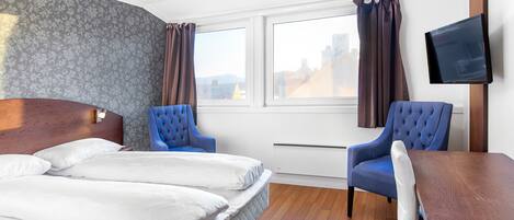 A hotel room with a double bed, two blue armchairs, a wooden desk, and a flat-screen TV mounted on the wall.