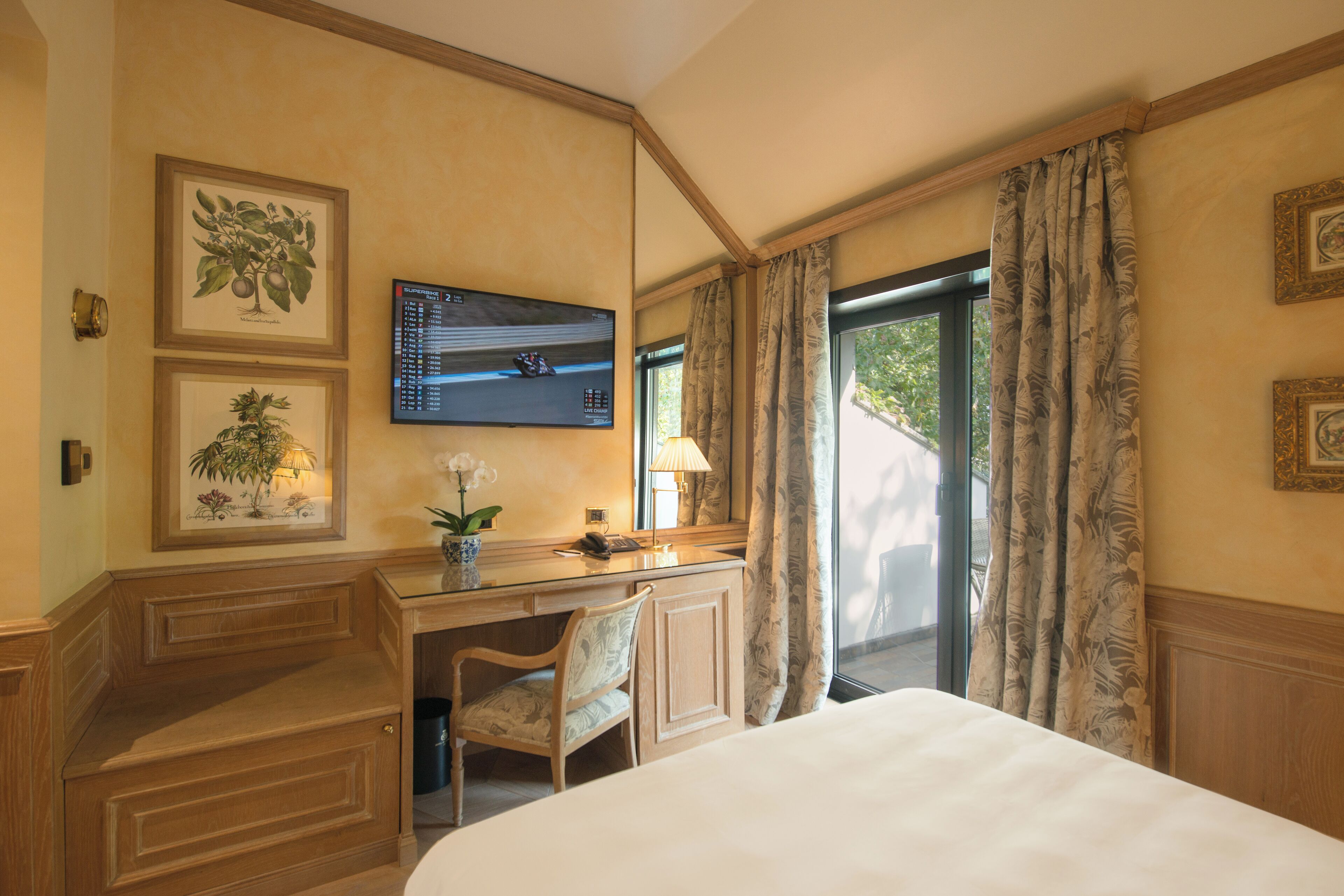 Habitación de hotel con cama, escritorio con silla, televisión e impresiones botánicas enmarcadas en la pared.