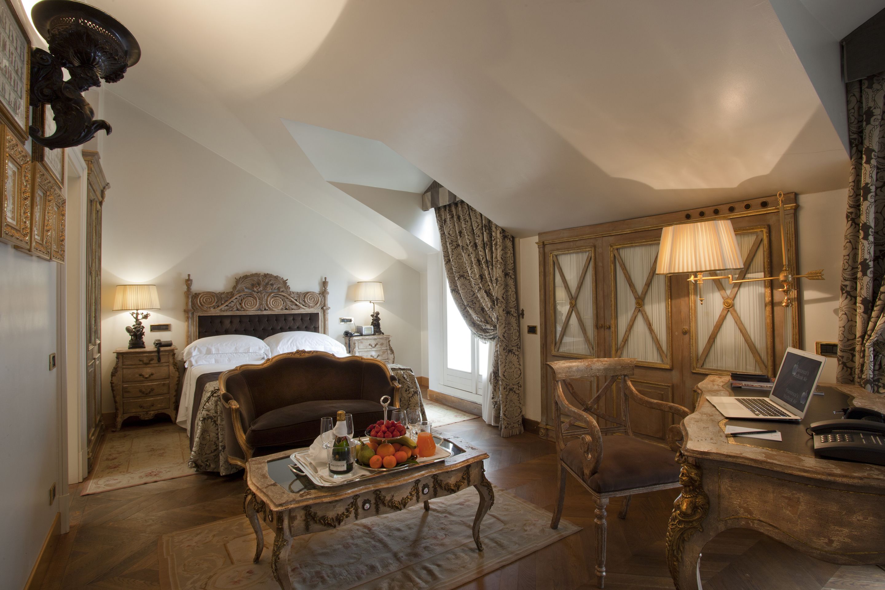 A bedroom with a bed, a desk with a laptop, a chair, a nightstand with lamps, and a tray with fruits and a bottle.