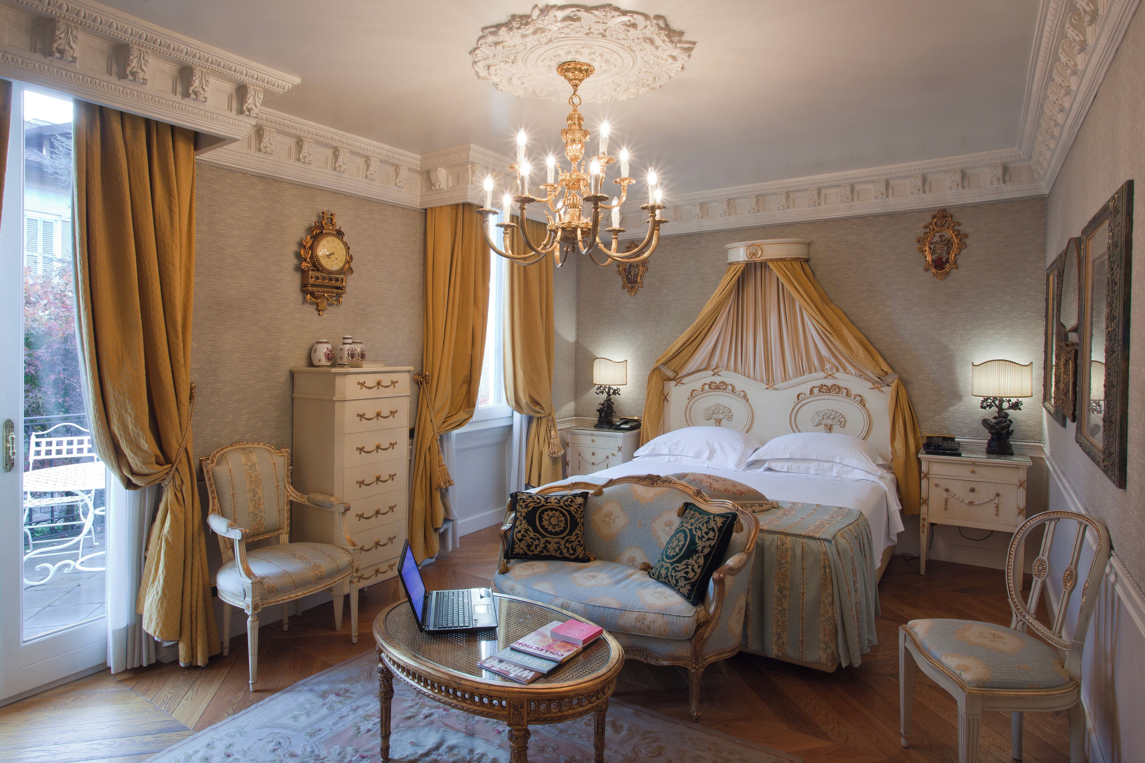 Um quarto luxuoso com uma cama de dossel, uma poltrona, um laptop sobre uma mesinha e uma mesa de cabeceira com abajur.