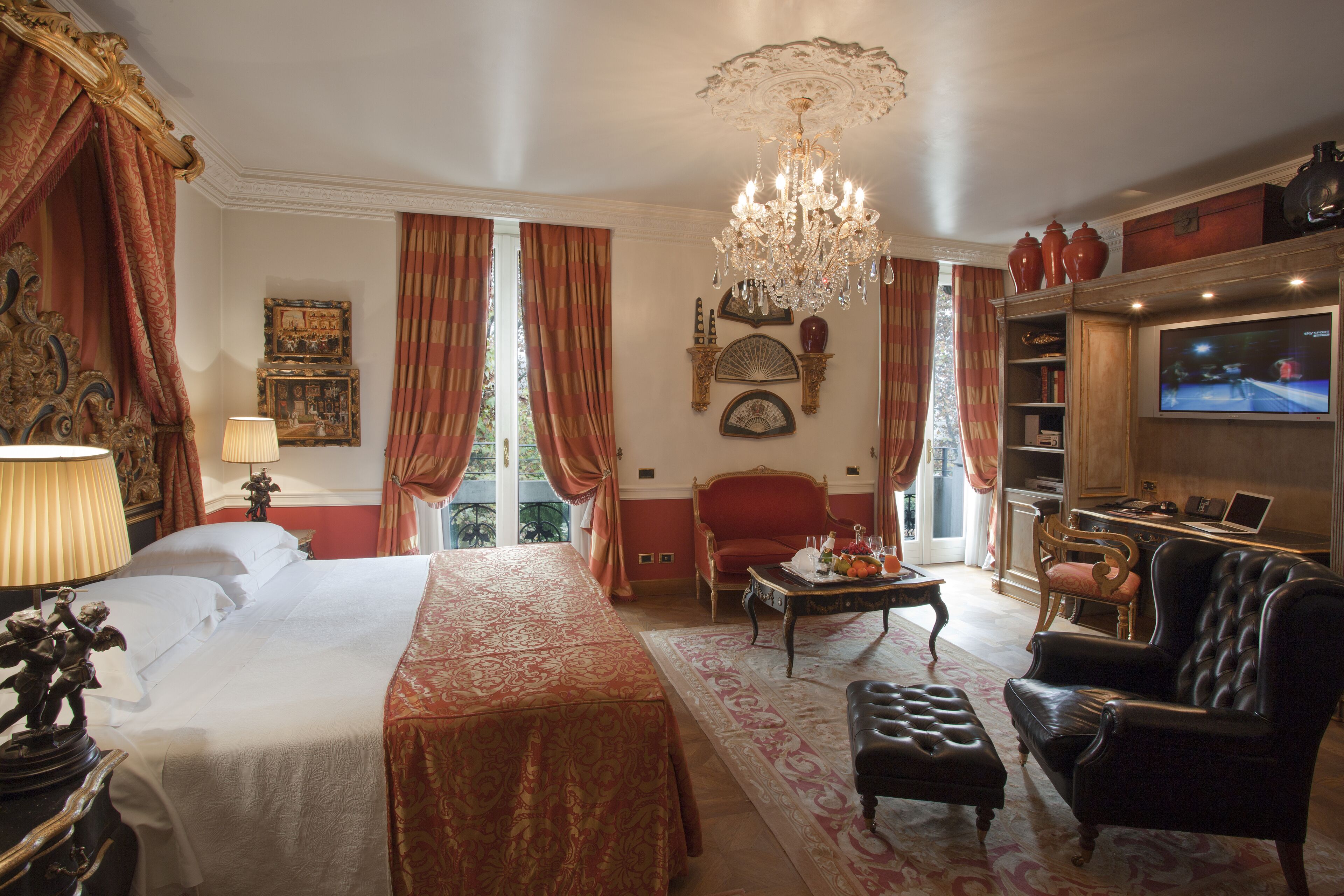 Habitación de hotel con una cama grande, una zona de estar con sofá y sillón, una araña y una televisión.