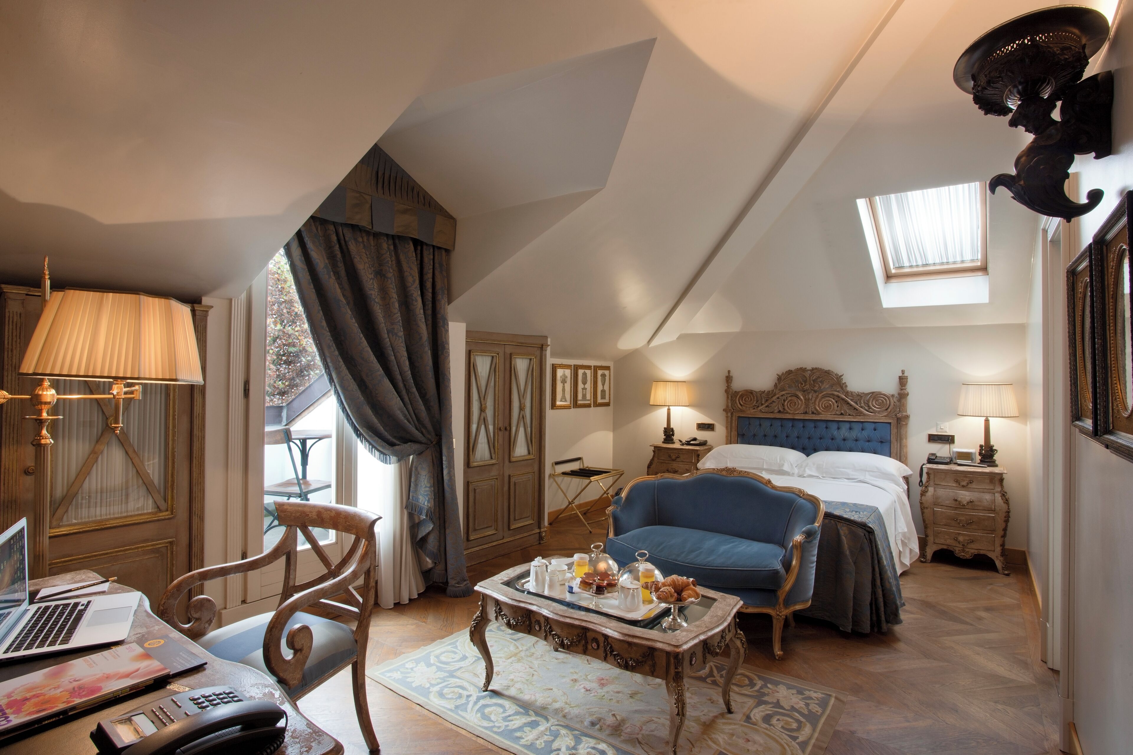 Ein Schlafzimmer mit Bett, Schreibtisch mit Laptop, Stuhl, Tisch mit Frühstücksgebäck und einem Fenster mit Vorhängen.