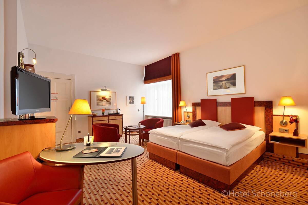 A hotel room with a bed, two bedside tables, a desk with a chair, a TV, and a window with curtains.