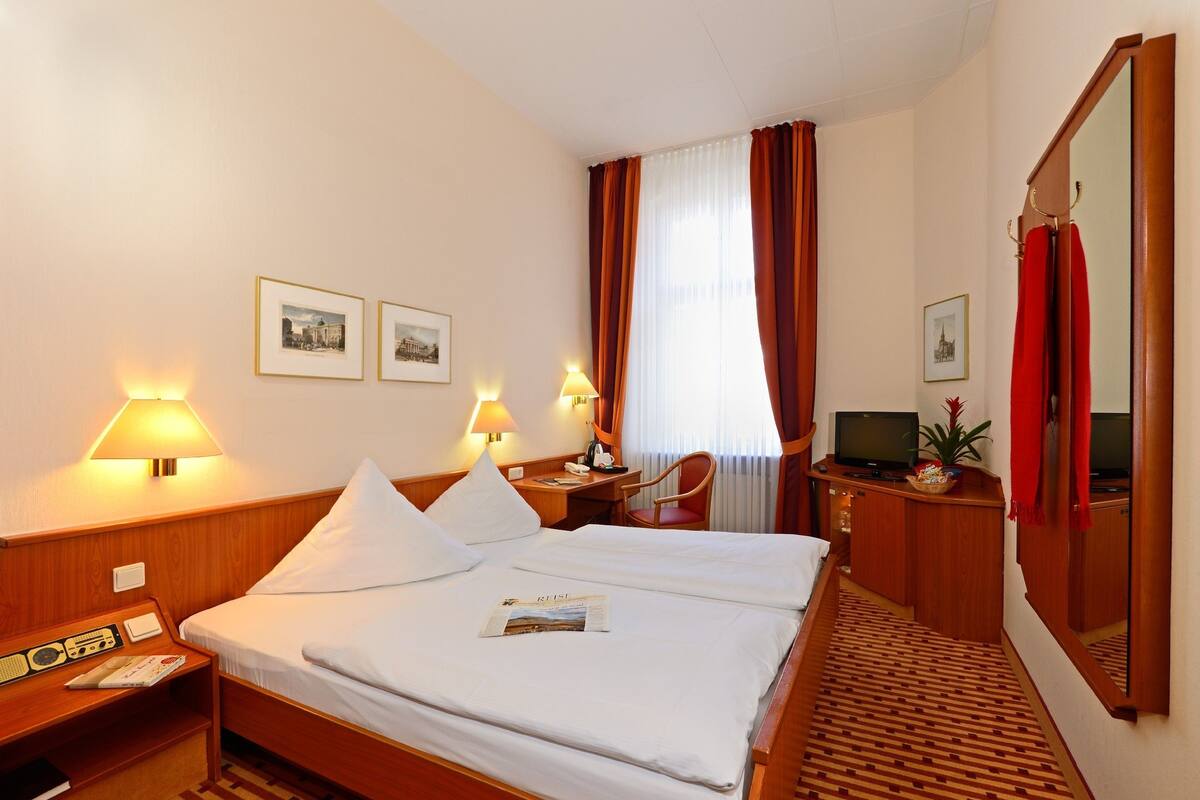 A hotel room with a bed, bedside table, lamp, chair, desk, TV, and a mirror.