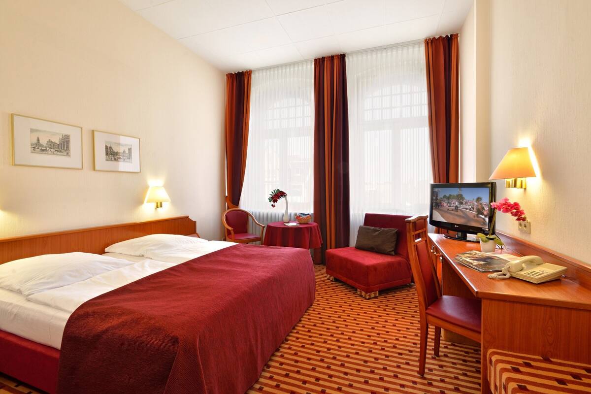 A hotel room with a large bed, a desk with a computer, a chair, a small table, and a TV.