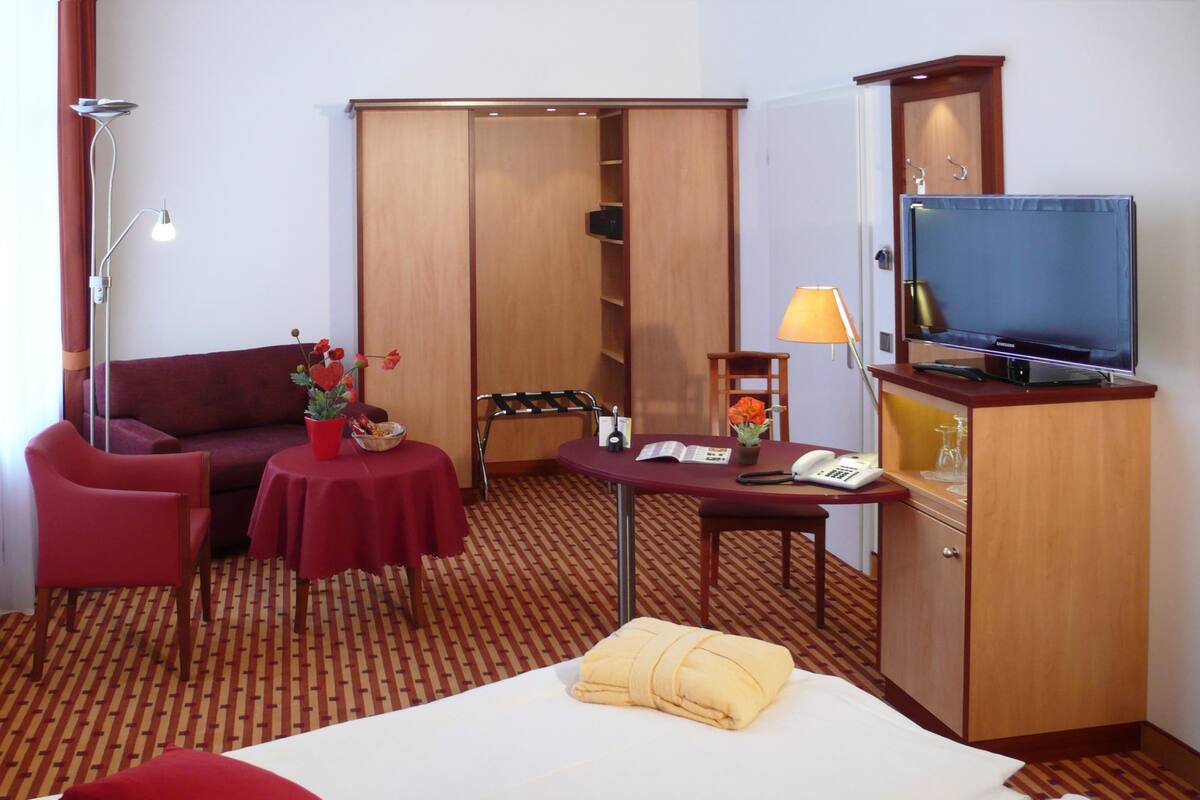 A hotel room with a bed, a desk with a telephone, a television, a sofa, a table with flowers, and a wardrobe.