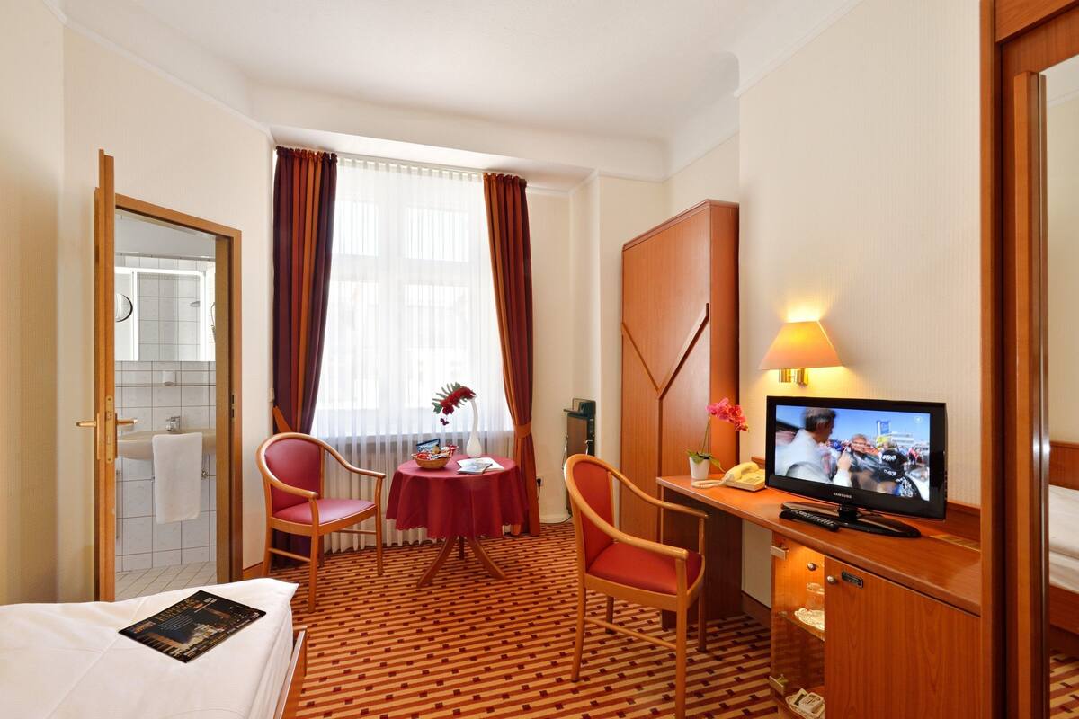 A hotel room with a bed, a desk with a television, a chair, a small table with a vase, and a window with curtains.