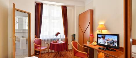 A hotel room with a bed, a desk with a television, a chair, a small table with a vase, and a window with curtains.