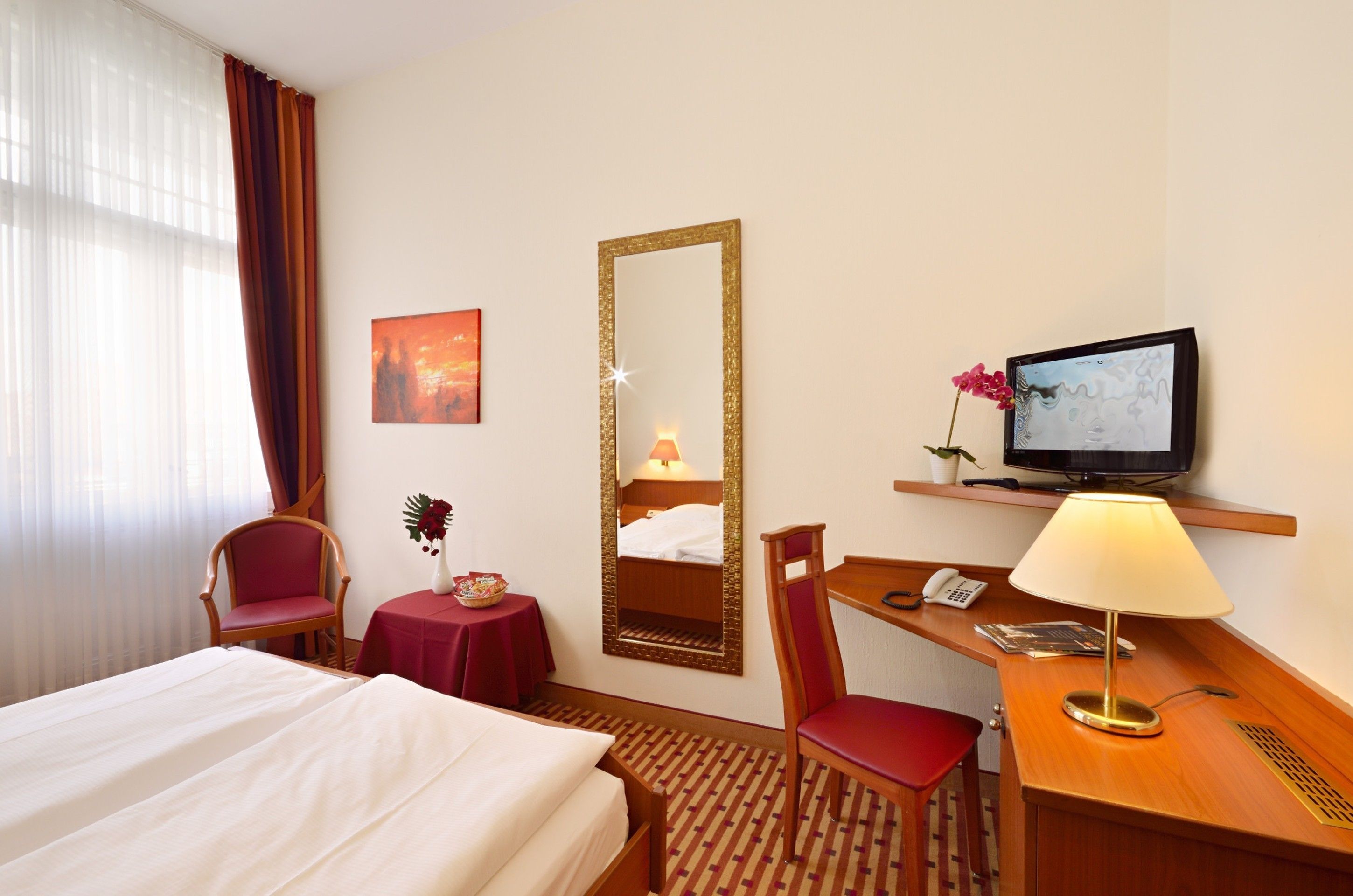 Habitación de hotel con cama, escritorio con lámpara, silla, televisor, espejo y una pequeña mesa con un jarrón de flores.