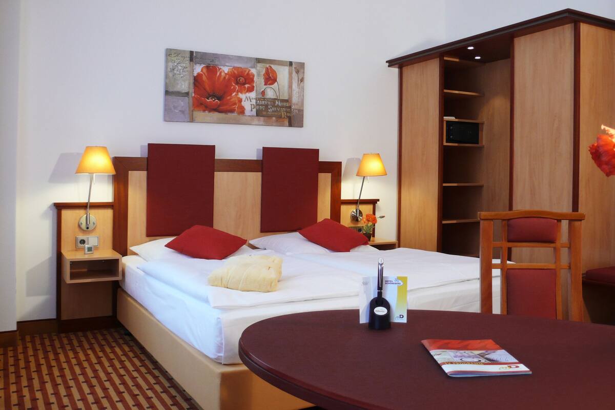 A hotel room with a bed, two bedside lamps, a nightstand, a wooden wardrobe, a desk with a chair, and a vase with flowers.