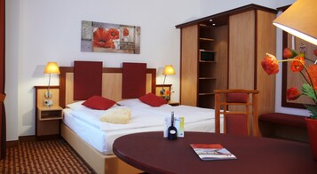 Una habitación de hotel con una cama, dos lámparas de noche, una mesita de noche, un armario de madera, un escritorio con una silla y un florero con flores.