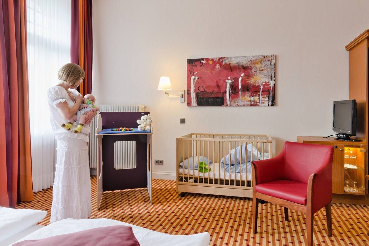 A hotel room with a crib, a red chair, a television, and a painting on the wall.
