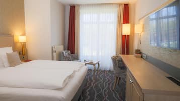 Ein Hotelzimmer mit einem großen Bett, einem Schreibtisch, einem Sessel, einem Heizkörper und einem Fenster mit Vorhängen.