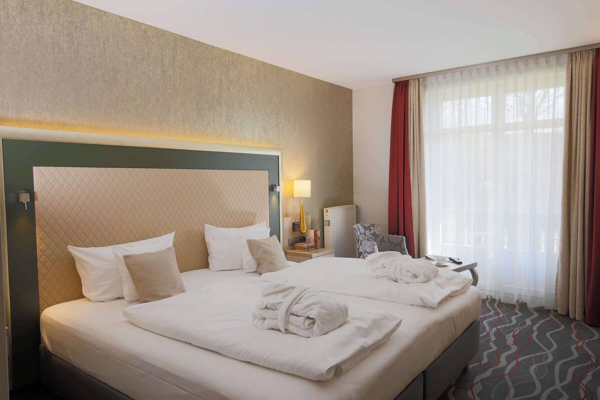 A hotel room with a large bed, a bedside table with a lamp, a chair, and a view through sheer curtains.