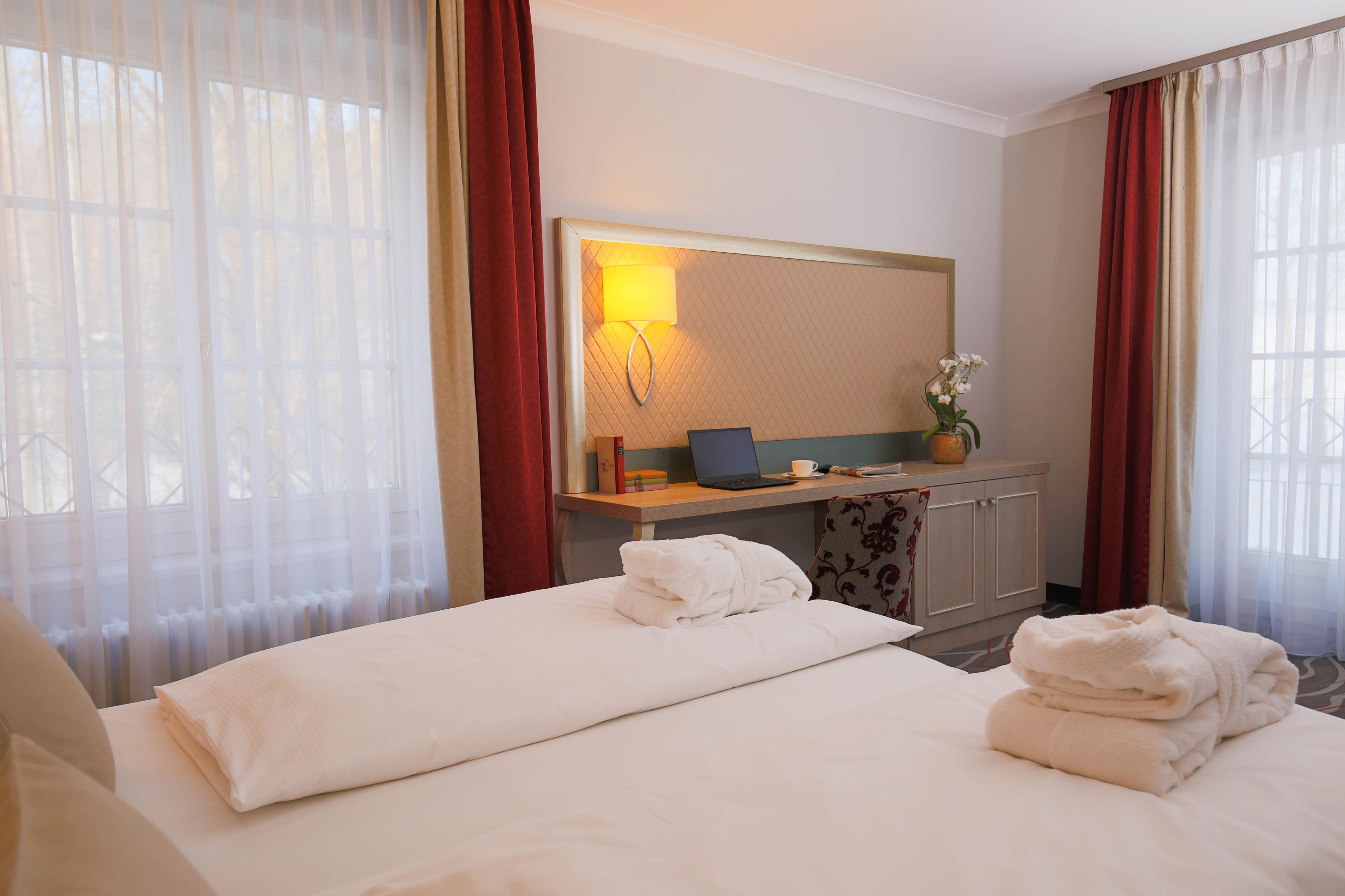 A hotel room with a bed, a desk with a laptop, a mirror, and a window with curtains.