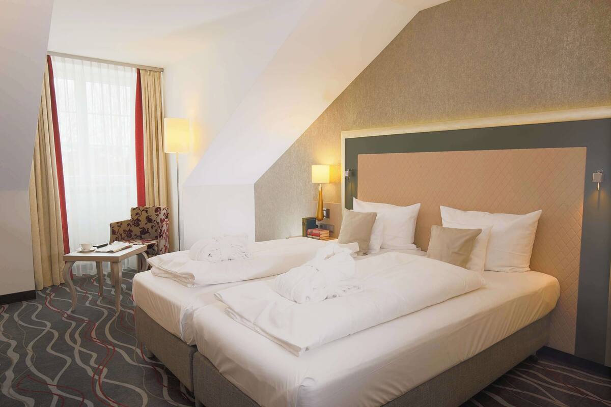 A hotel room with two beds, a desk, a chair, and a lamp.