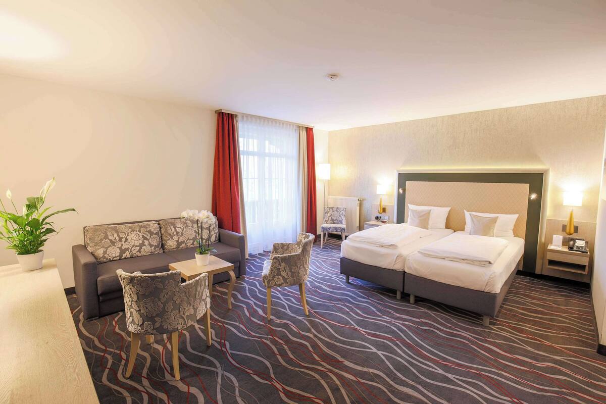 A hotel room with a bed, a sofa, a chair, a table, and a potted plant.